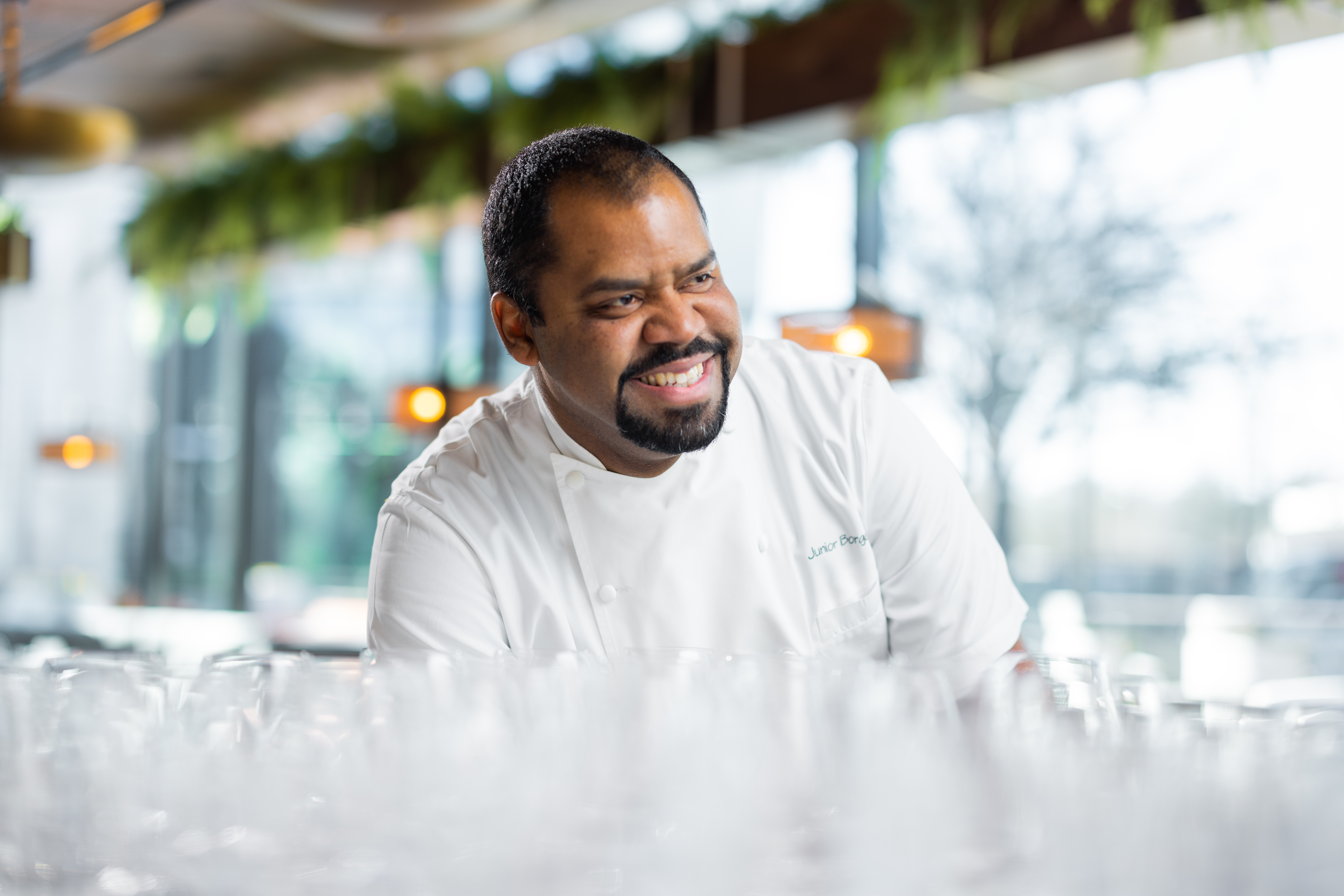 A chef in his whites leans over a counter. Out of focus in front of him are several glasses. He looks to the right and smiles.