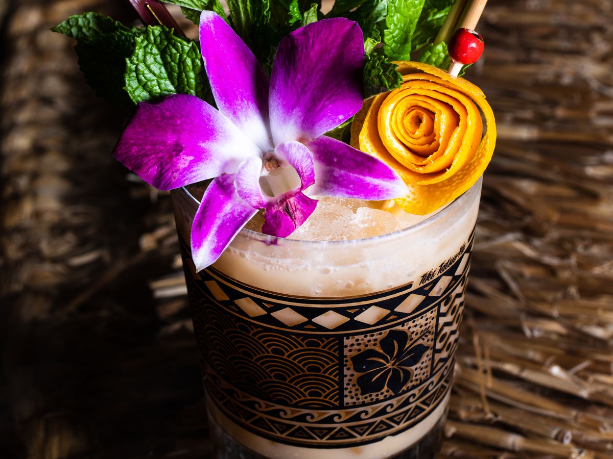 Cocktail topped with an orchid, green mint, and a mango peel curled into a flower, with a pineapple-shaped skewer decoration.