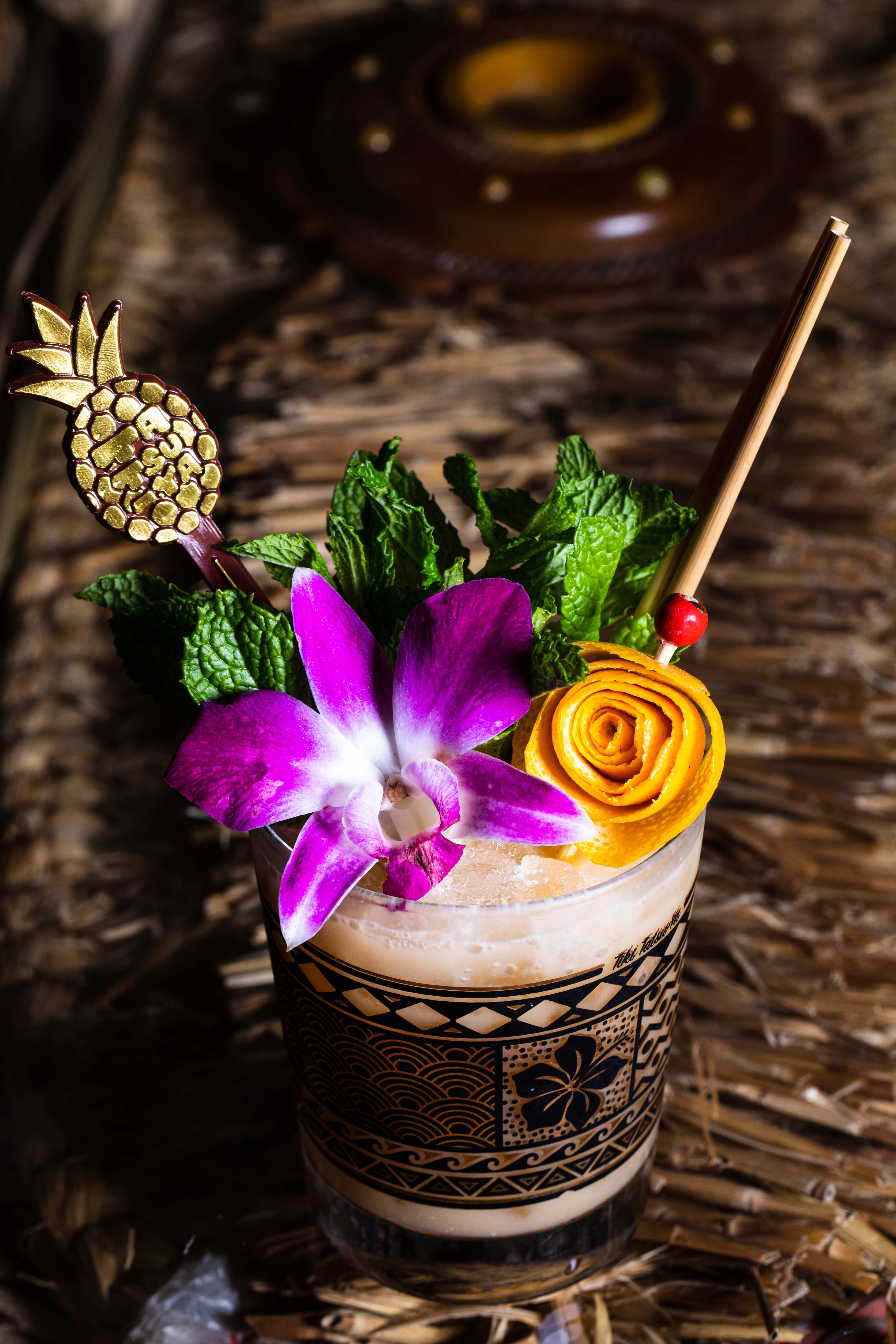 Cocktail topped with an orchid, green mint, and a mango peel curled into a flower, with a pineapple-shaped skewer decoration.