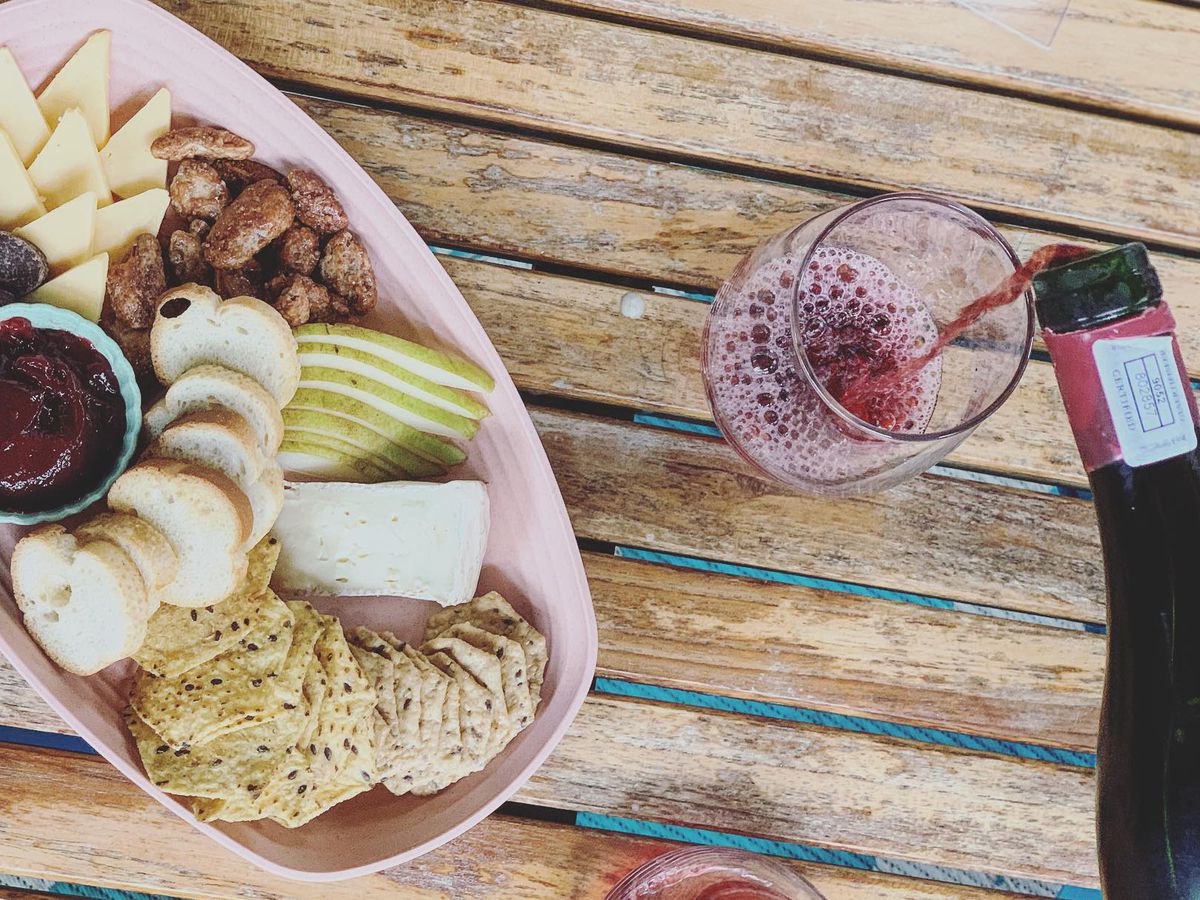 A plate of sliced cheeses, breads, and fruits next to someone pouring a glass of red wine.