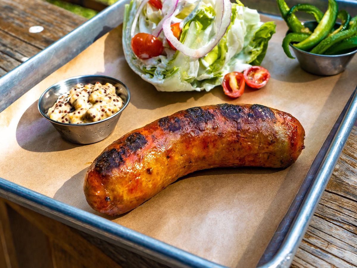 A sausage on a tray with a metal container of mustard next to a salad.
