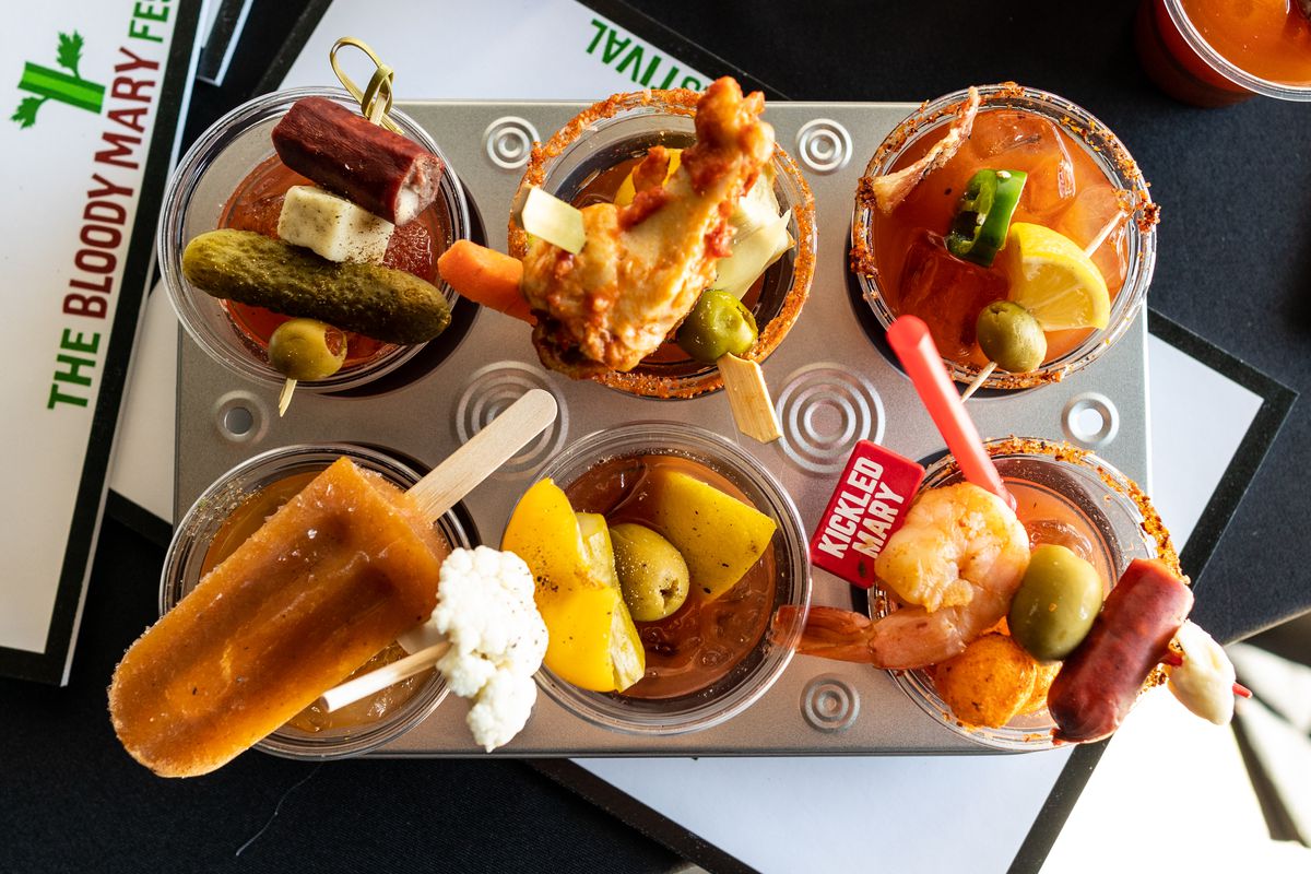 A half dozen mini-Bloody Marys, all garnished with different toppings, sit in a drinks container.
