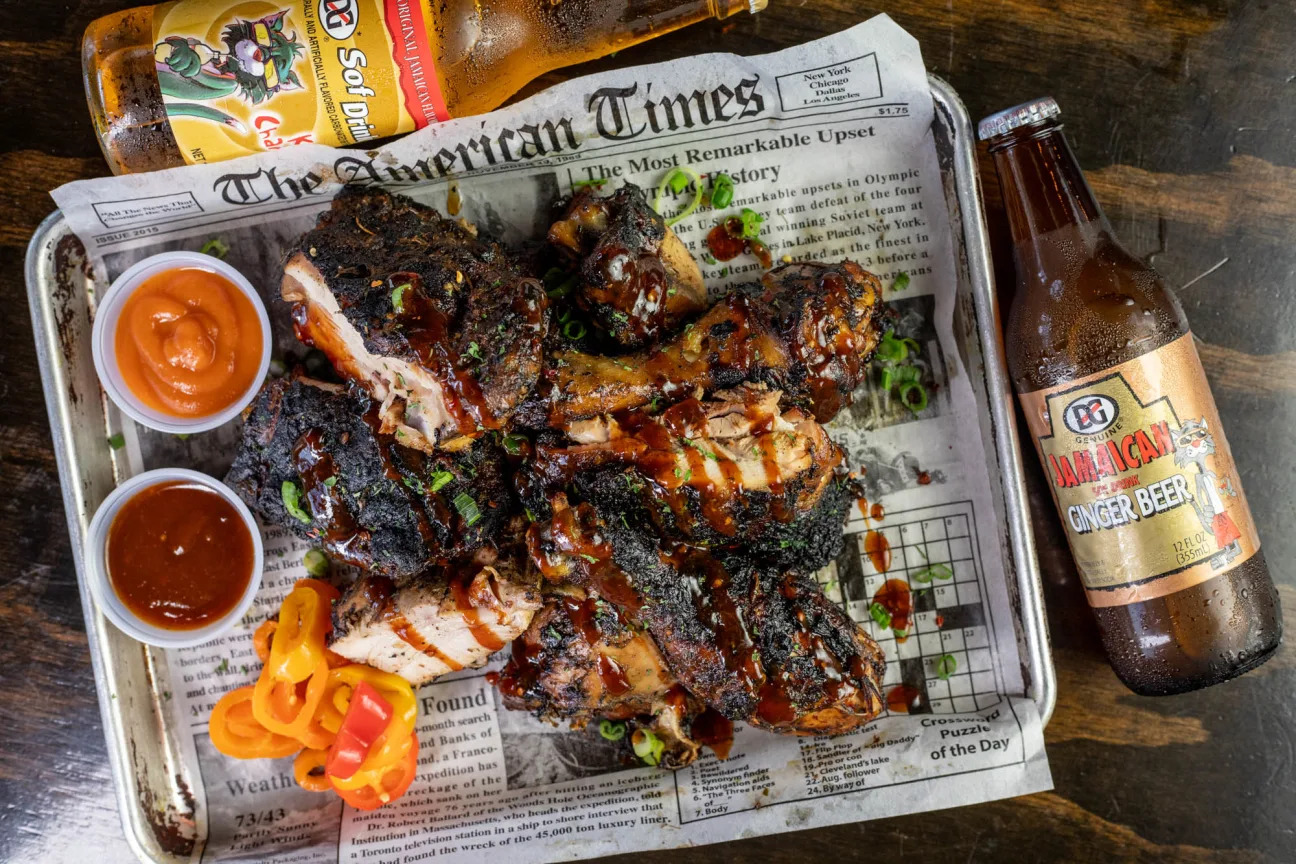 A newspaper-lined tray of charred saucy chicken with sides. 