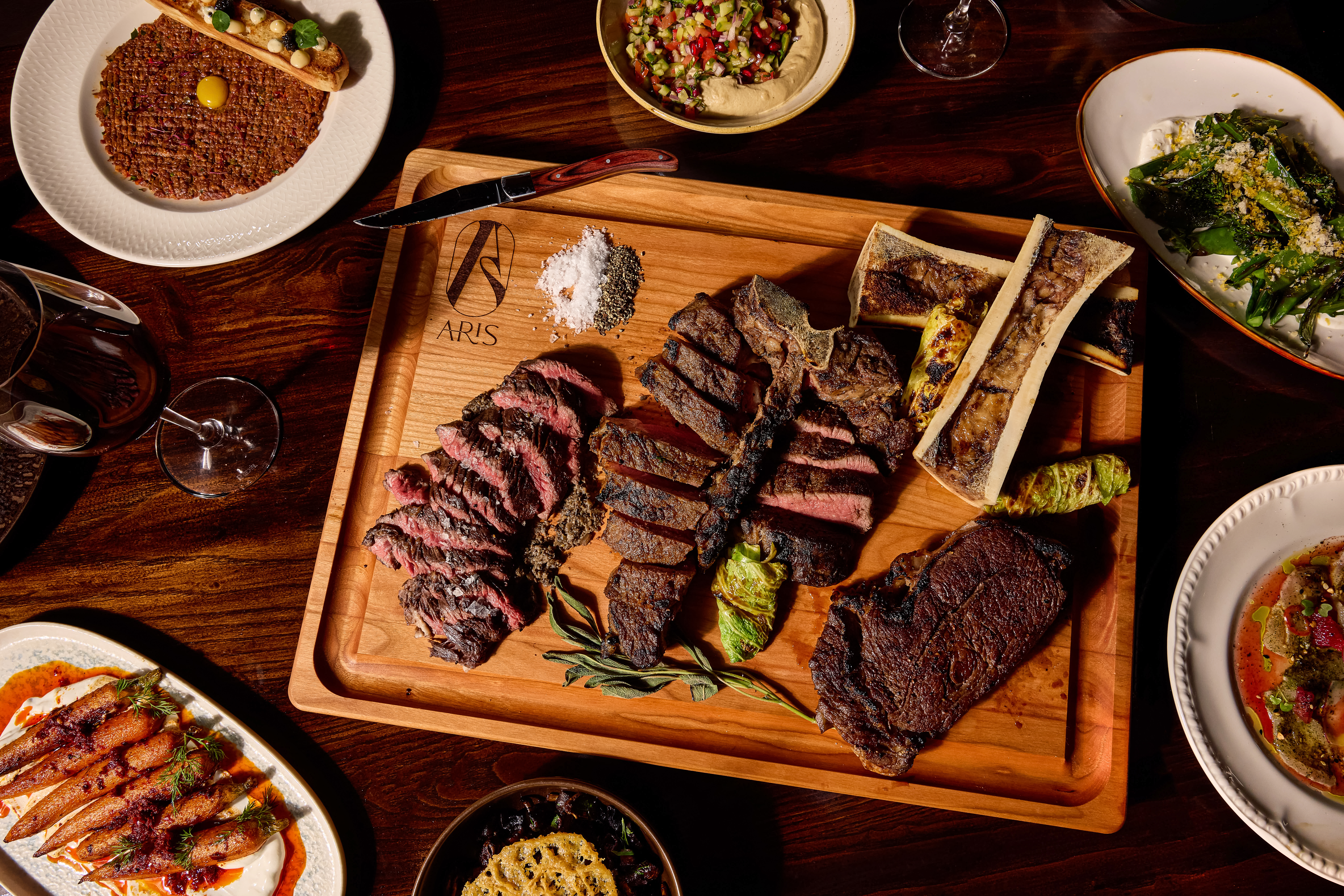 A spread of Aris dishes, including a sliced steak, baked carrots, and fries.