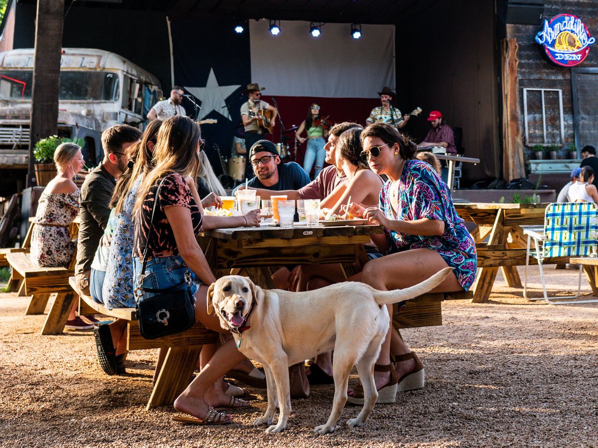 A group sits outside at a picnic table at a bar, with a Texas flag in the background. A woman in the foreground holds the collar of a golden lab.