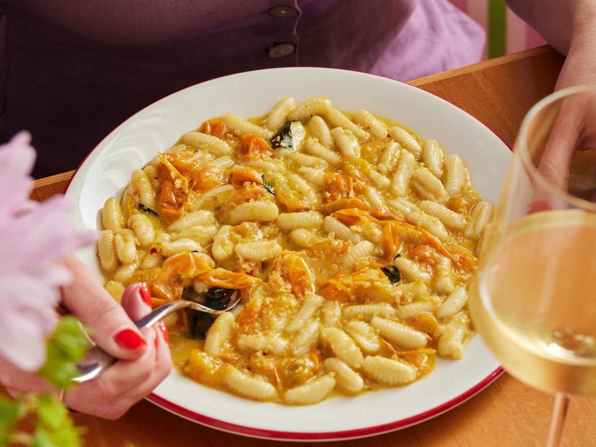 A plate of pasta is on a table with a glass of white wine. A woman sticks a spoon into it to eat.