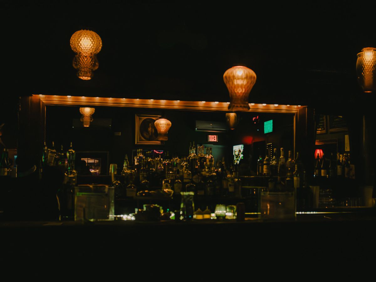 The interior of a bar that’s extremely dark shows the bar back, hanging lights, and bottles of liquor.