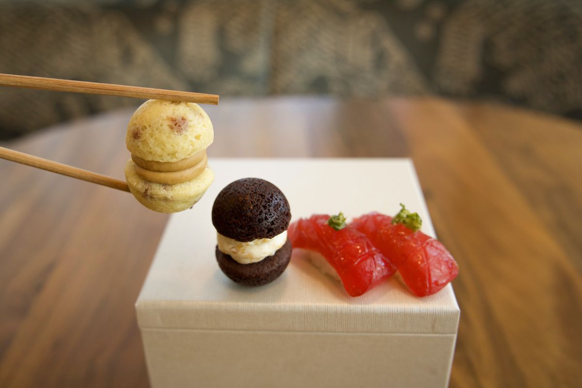 A pair of chopsticks hold a mini white chocolate macaroon while chocolate version sits on a box next to it, with two pieces of sushi nearby.