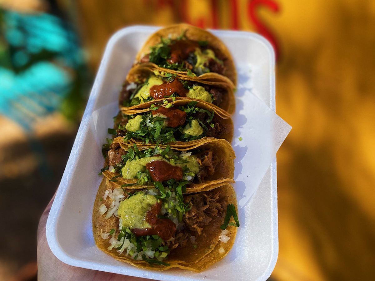 A hand holds up a white tray of a half dozen street tacos topped with salsa verde and rojas.