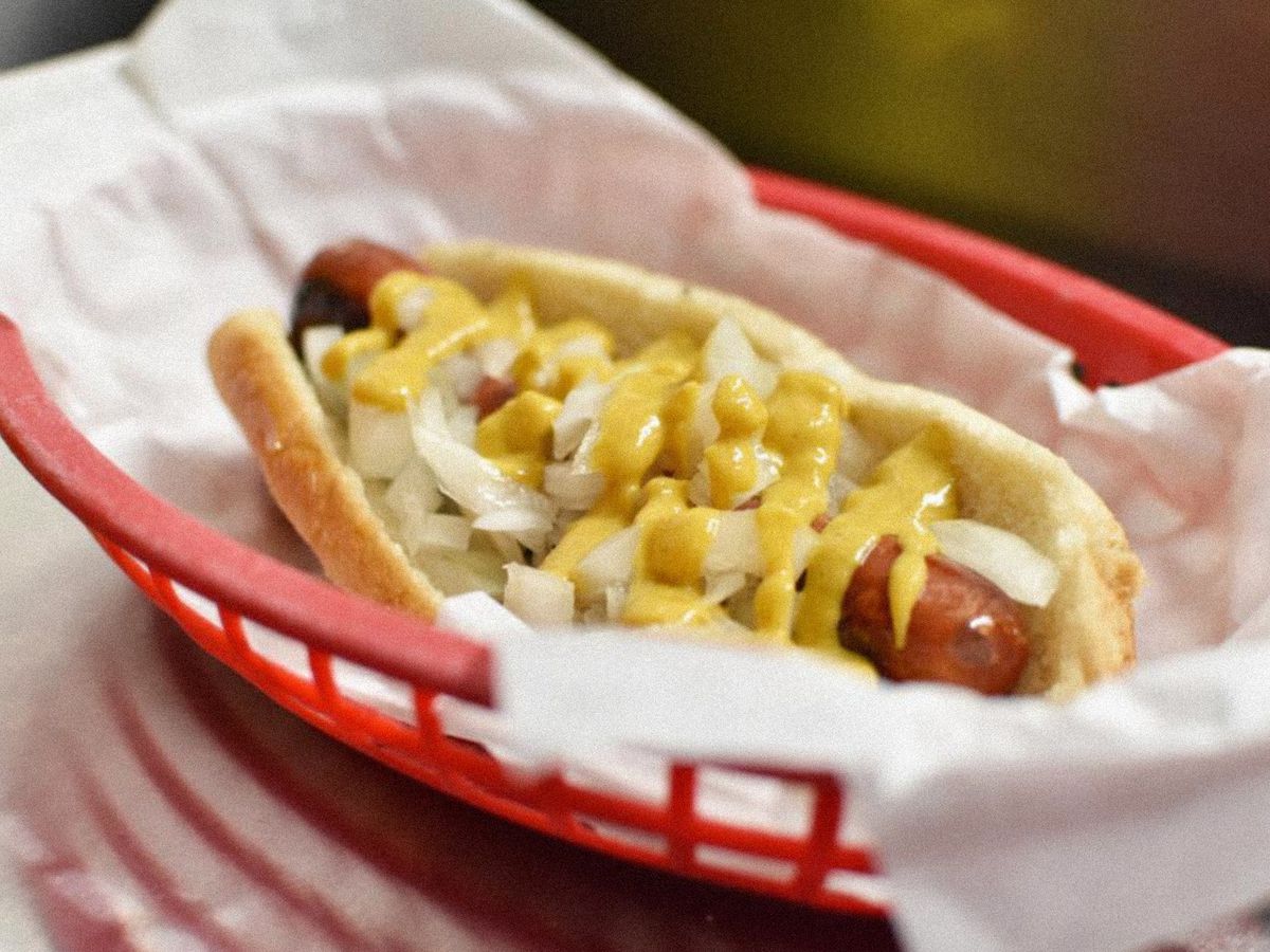 A hot dog sits in a red basket lined with white paper. It is topped with melted cheese and onions.