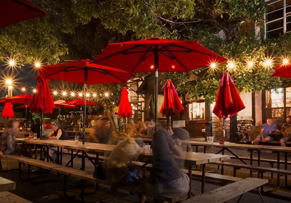 A time lapse photo shows people moving around at an outside beer garden at night, with red umbrellas above the tables.