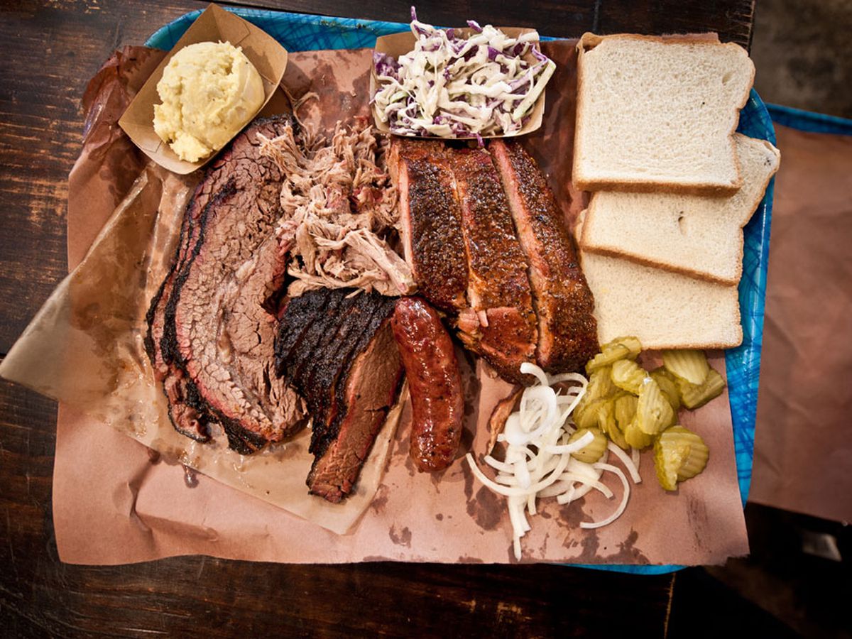 A tray of various barbecue meats ranging from brisket slices to ribs to sausages, alongside sides of coleslaw and potato salad and slices of bread, pickle slices, and onions.