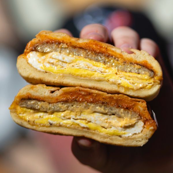A hand holds two sides of an open egg, sausage, and cheese sandwich up to the camera.