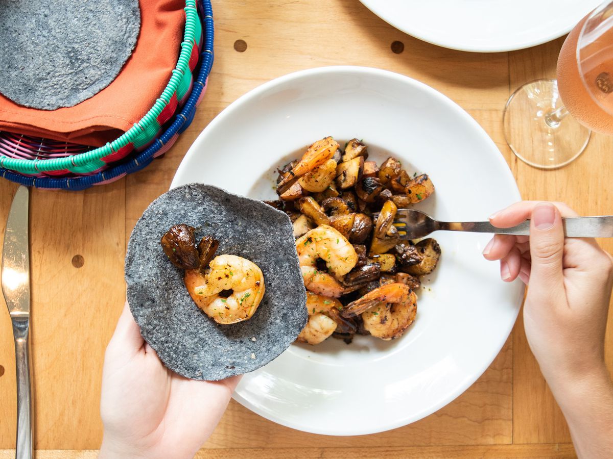 A person holding up a blue tortilla with a shrimp on it with one hand and spooning a bowl of shrimp with the other