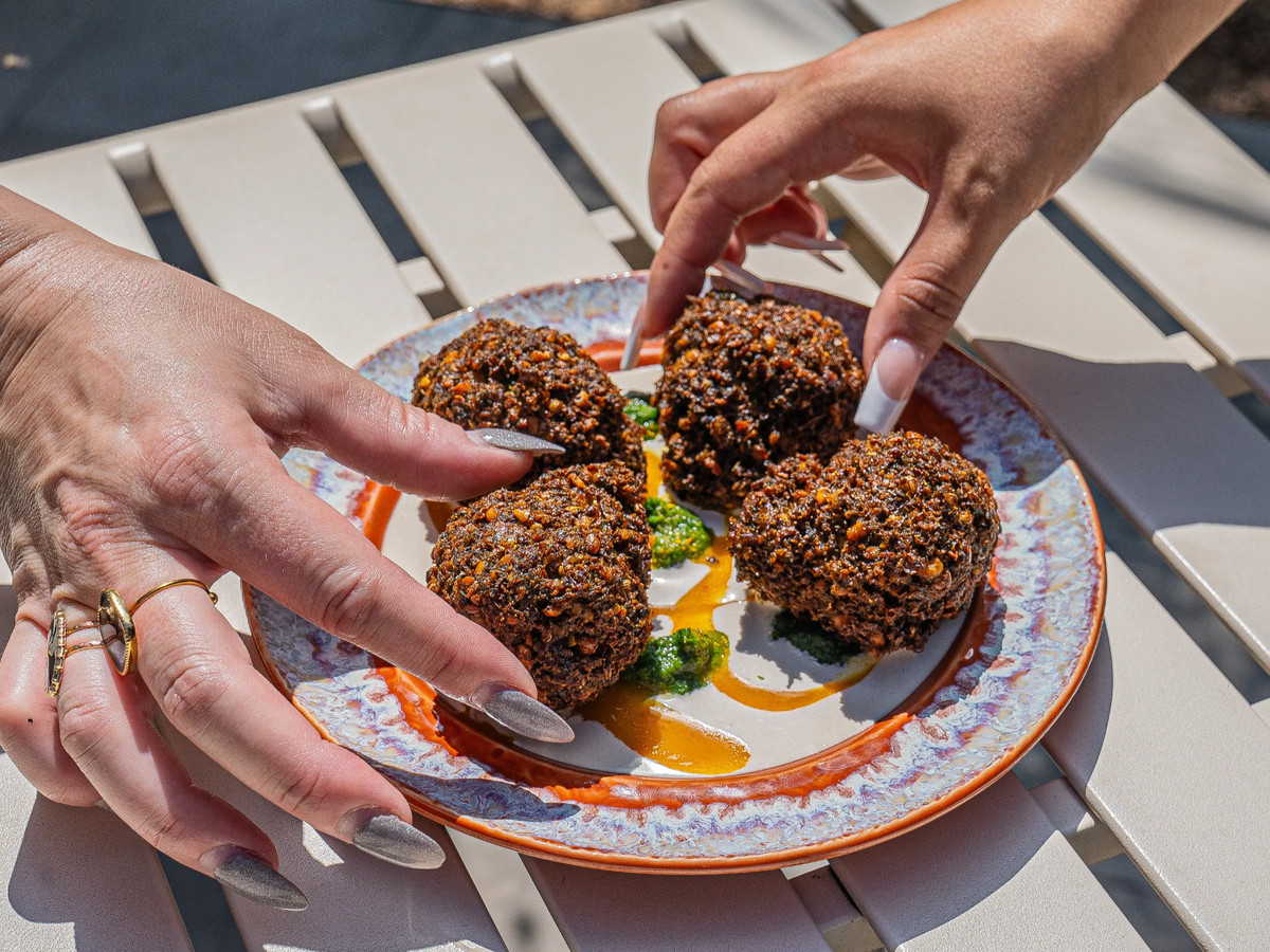 A plate holds four pieces of falafel. The hands of two women reach in to grab one each.