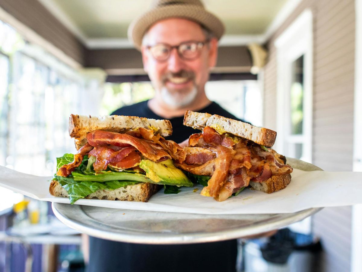 A man holds a plate with a BLT on it, which he thrusts towards the camera.