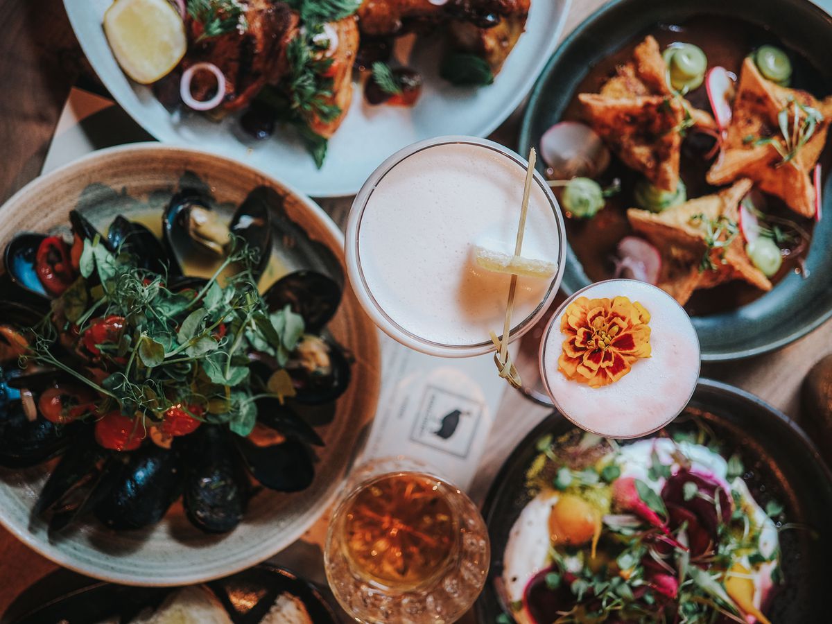 A table holds a trio of cocktails in the middle of several plates of food, including mussels, wontons, and crudo.