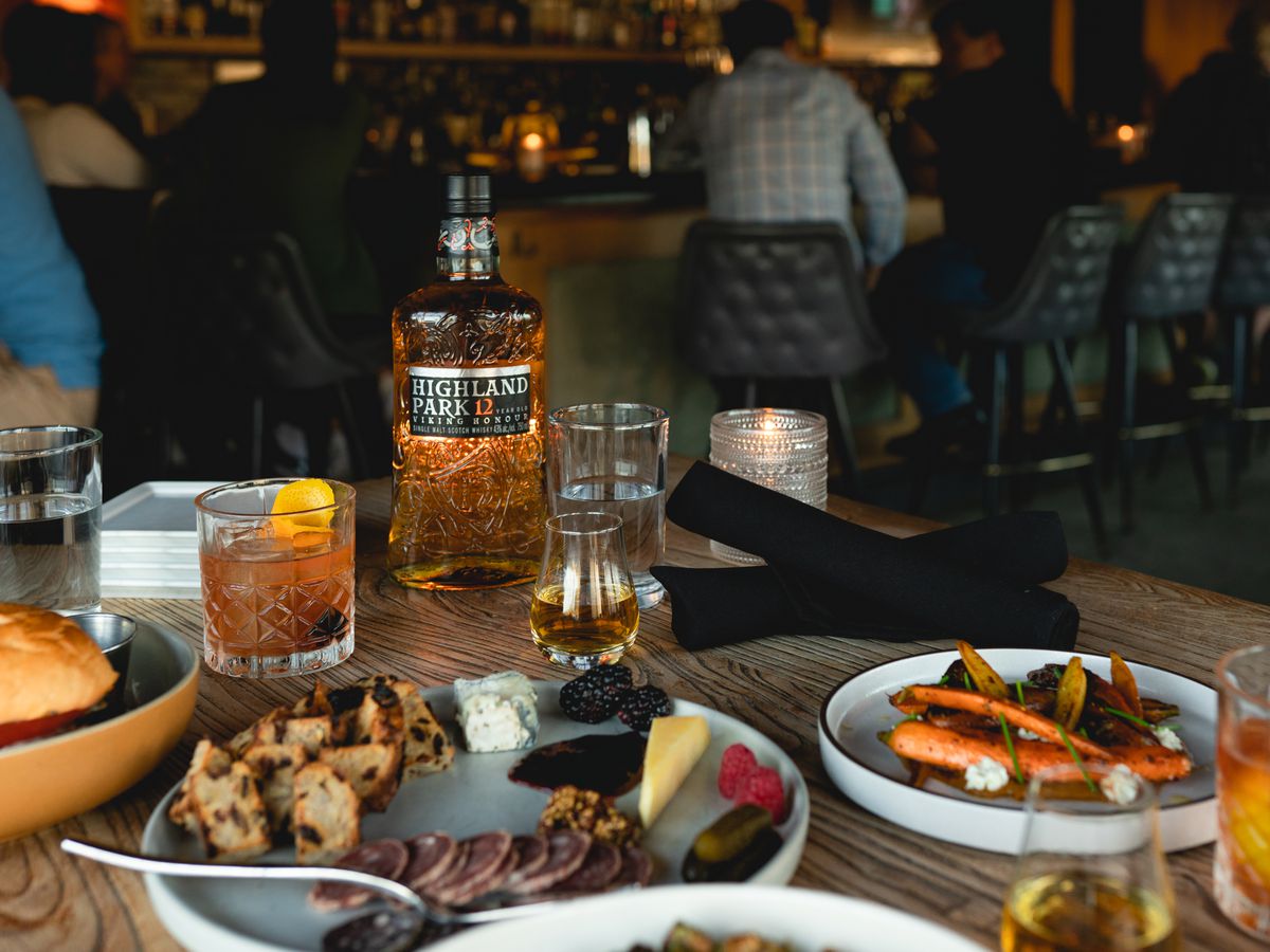A bottle of Highland Park whisky on a table next to cocktails and plates of charcuterie and sandwiches.