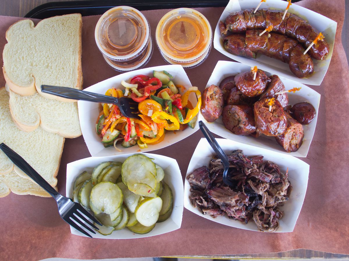 Several white cardboard trays of meats and pickles and peppers with slices of white bread on butcher paper.