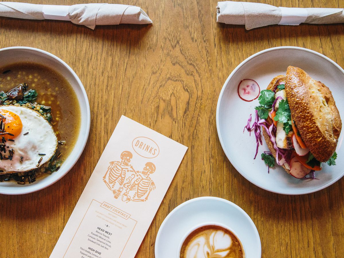 Two plates of food, one with a sunny-side-up egg and a broth and the other with a sandwich with a cup of latte and a drinks menu on a wooden table.
