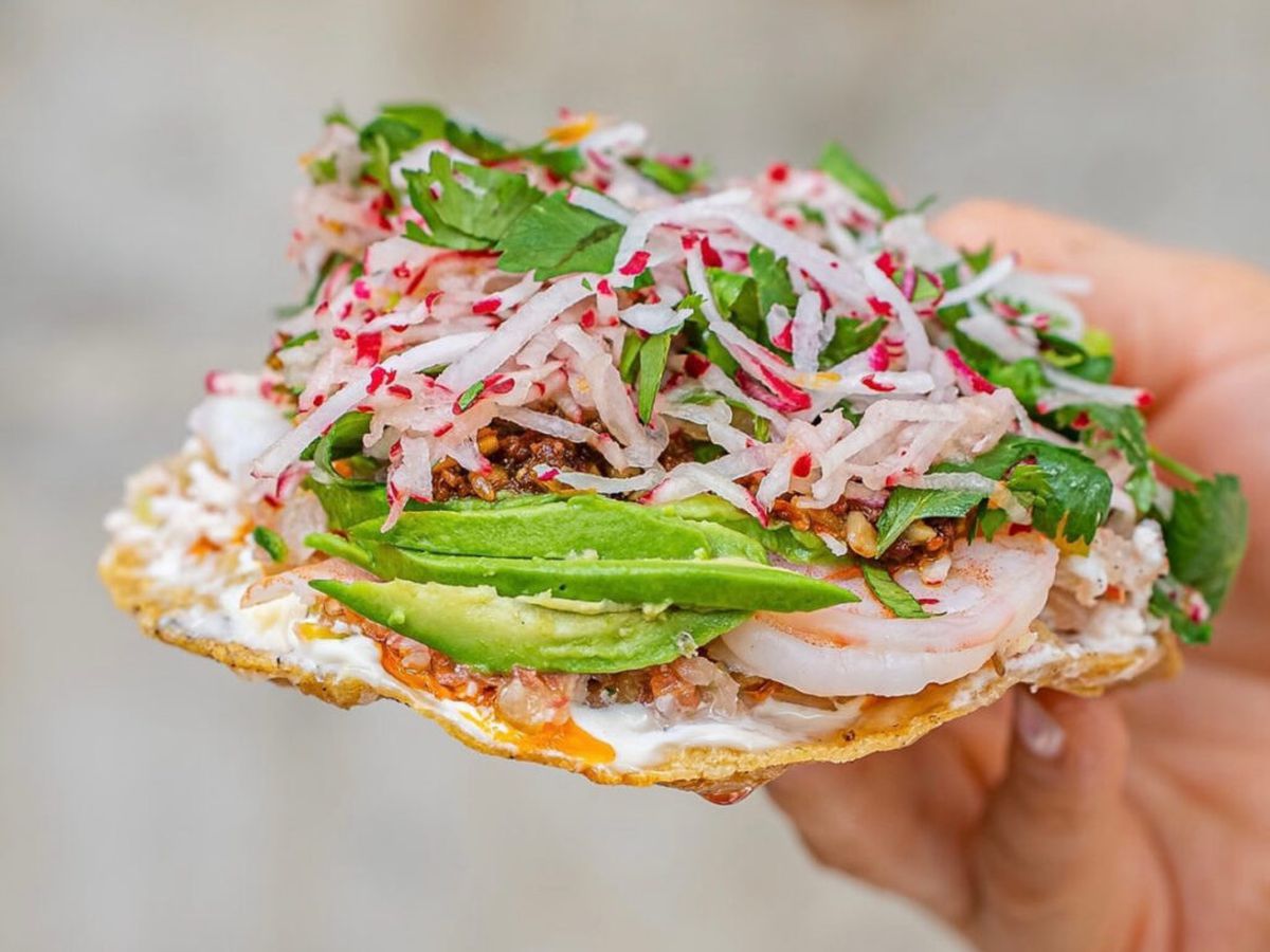 Someone holding up a tostada with avocado, shrimp, onions, and more.