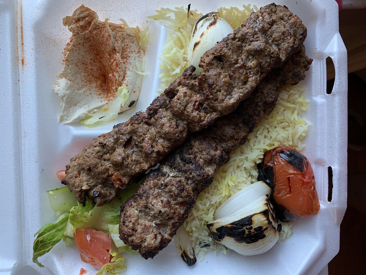 A container of yellow rice, grilled vegetables, a salad, hummus with spices, and two long meat kebabs.