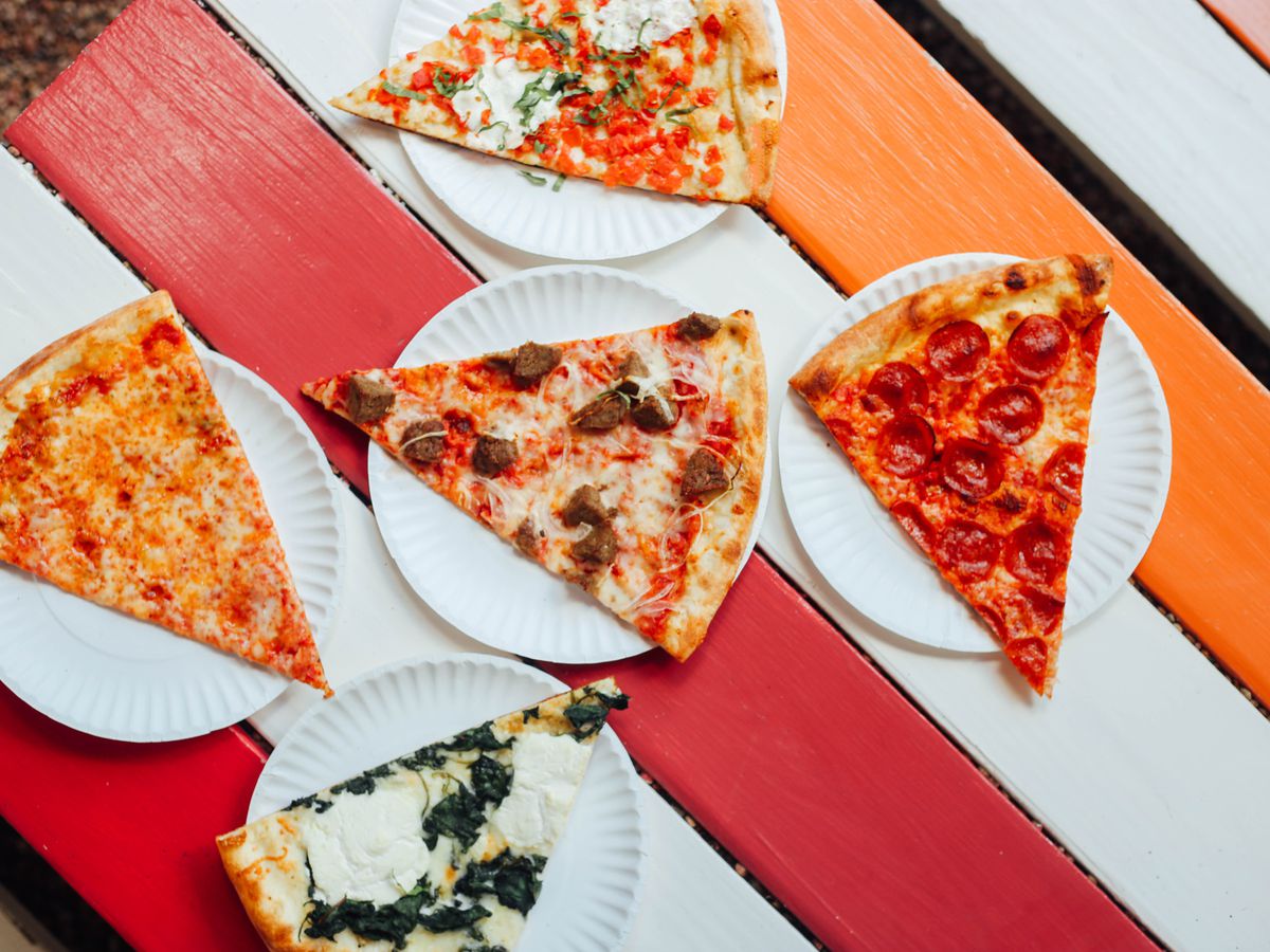 A bunch of pizza slices with various toppings on paper plates on top of a red-white-orange picnic table.