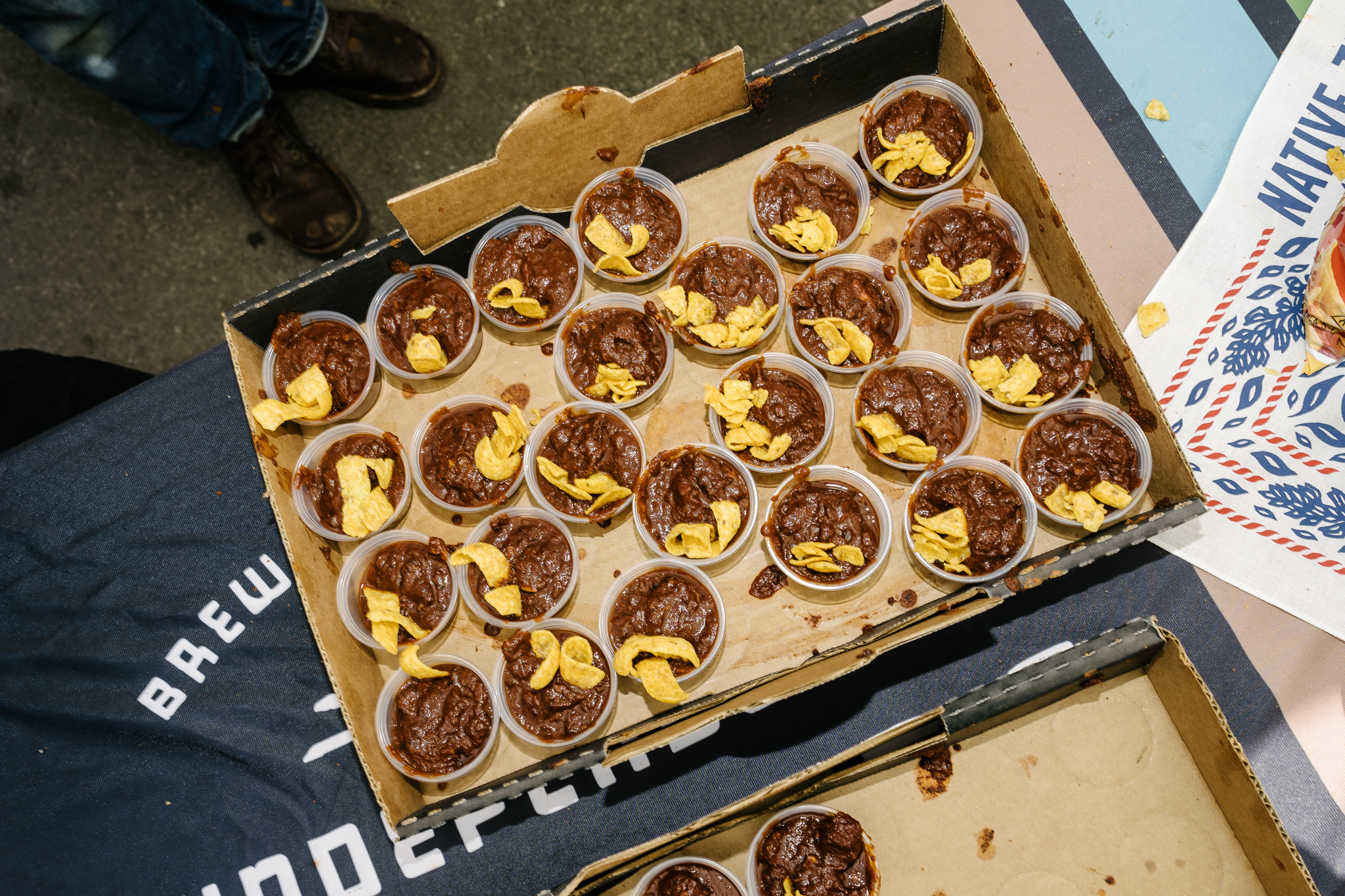 Several small taster sized containers of chili sit in a cardboard container with a few Fritos on top.