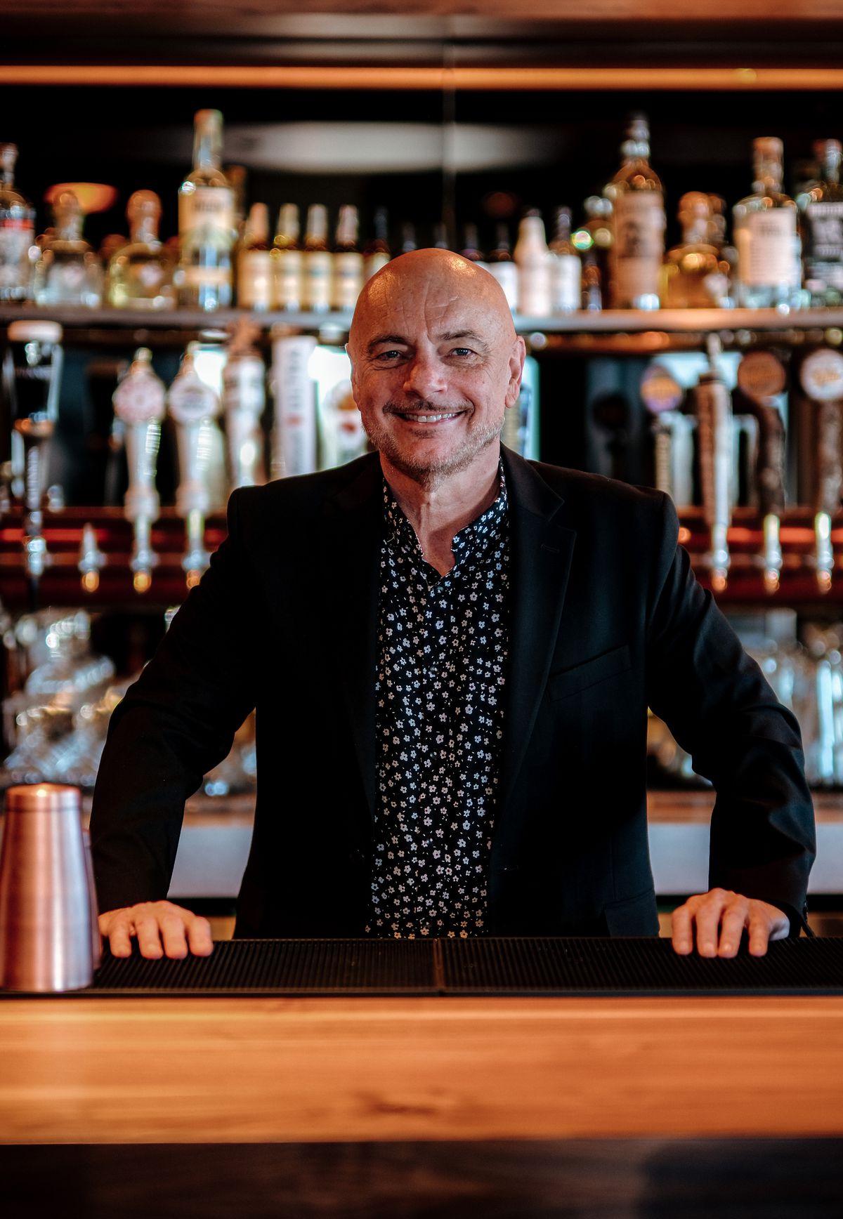 Food Network host John Green stands behind a bar in a black blazer and black shirt with white polk-a-dots.