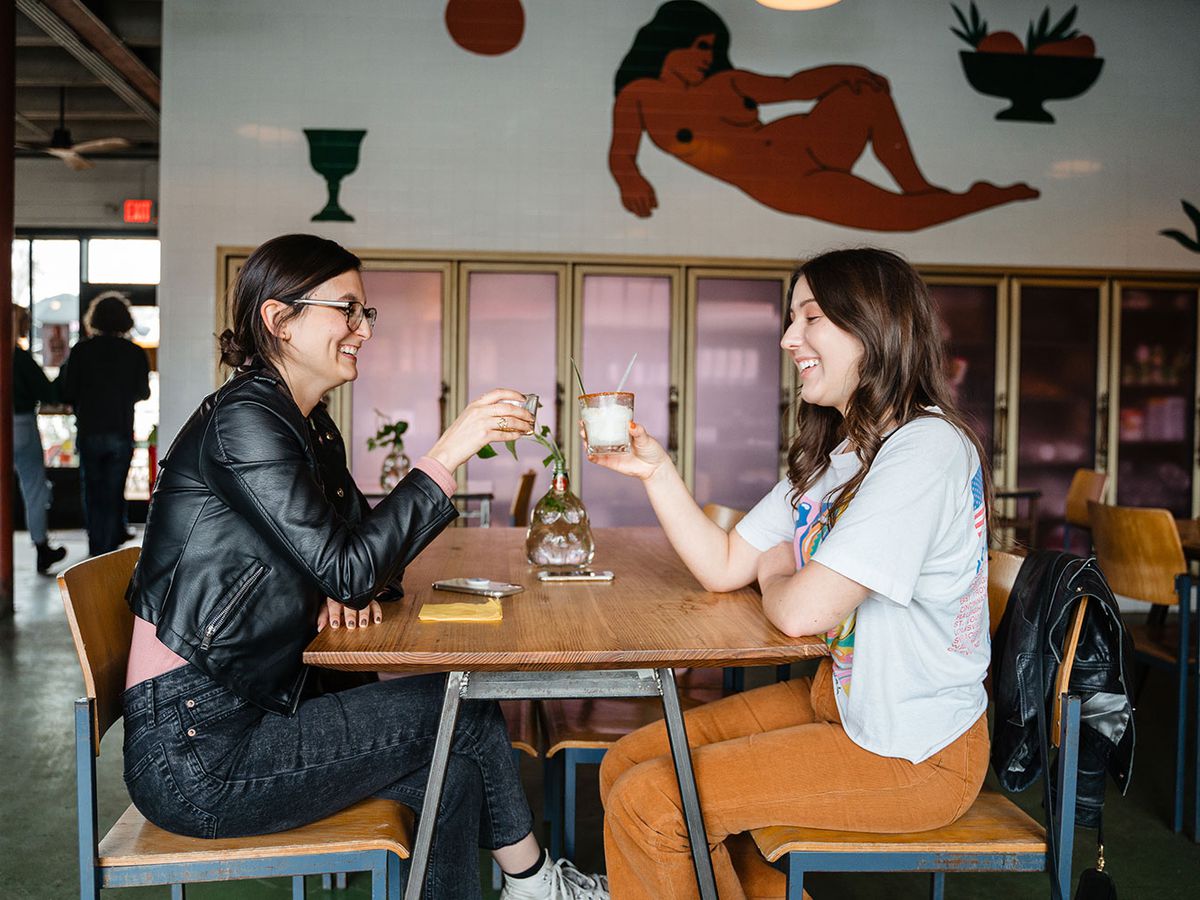 Two women sit at a table in a bar and raise their glasses in a cheers.