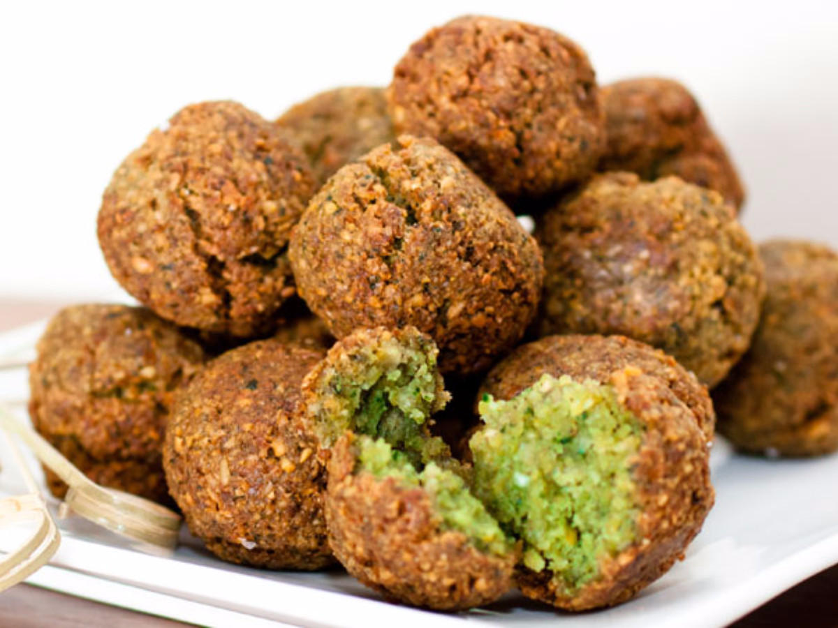 Balls of falafel are stacked on a plate. Near the bottom, one is open and green insides are shown.