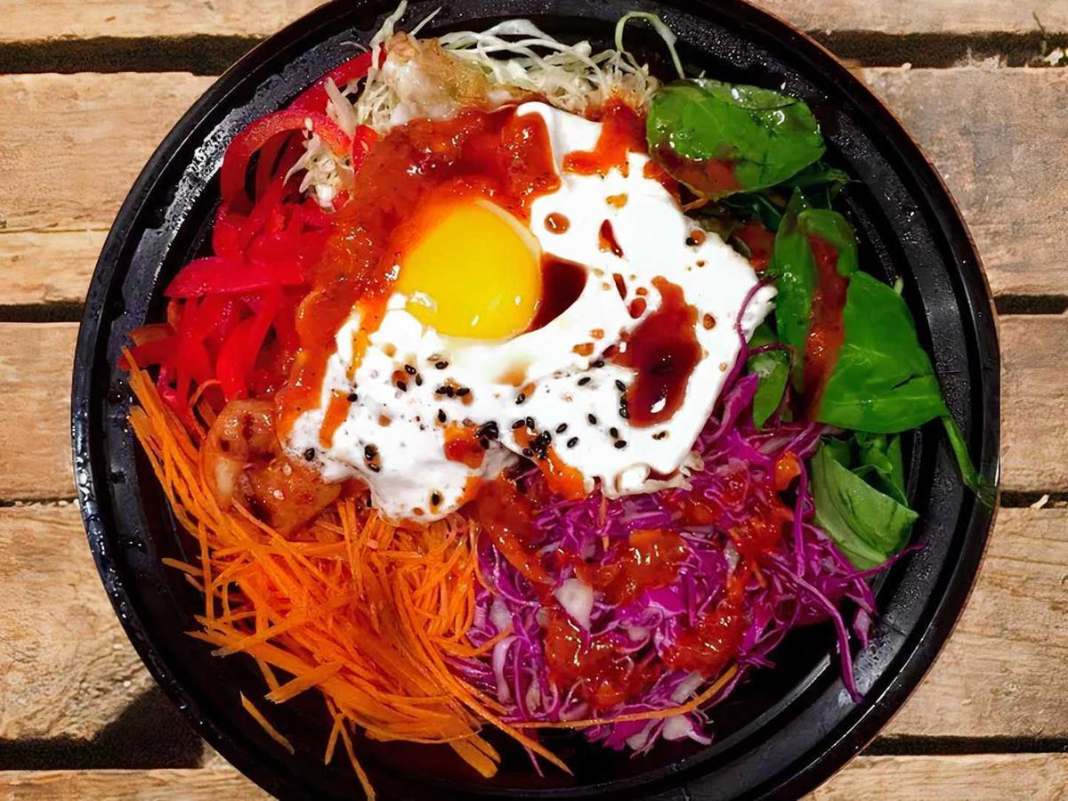 A bimbap bowl is topped with a soft fried egg, sliced carrots, red cabbage, and hot sauce.