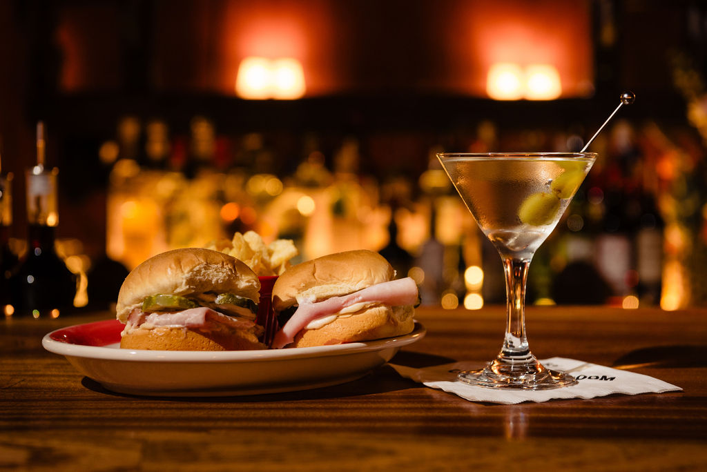 A table with a plate of two meat sandwiches next to a martini cocktail. 