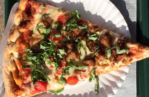 A triangle shaped slice of pizza sits on a paper plate. It is topped with sausage, tomatoes, and shredded basil.