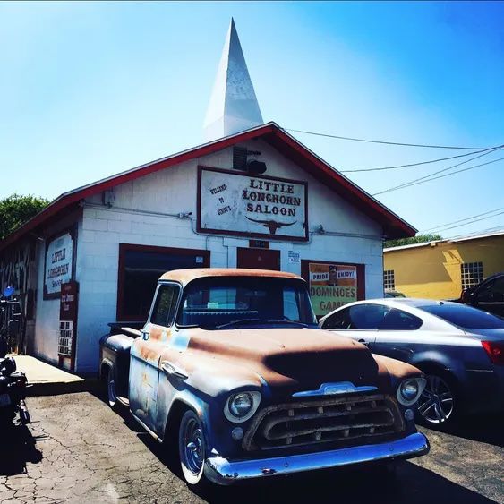 A white shack that has been converted to a bar has a sign out front that read “Little Longhorn Saloon.” A rusted out vintage truck is parked out front, next to a new car.