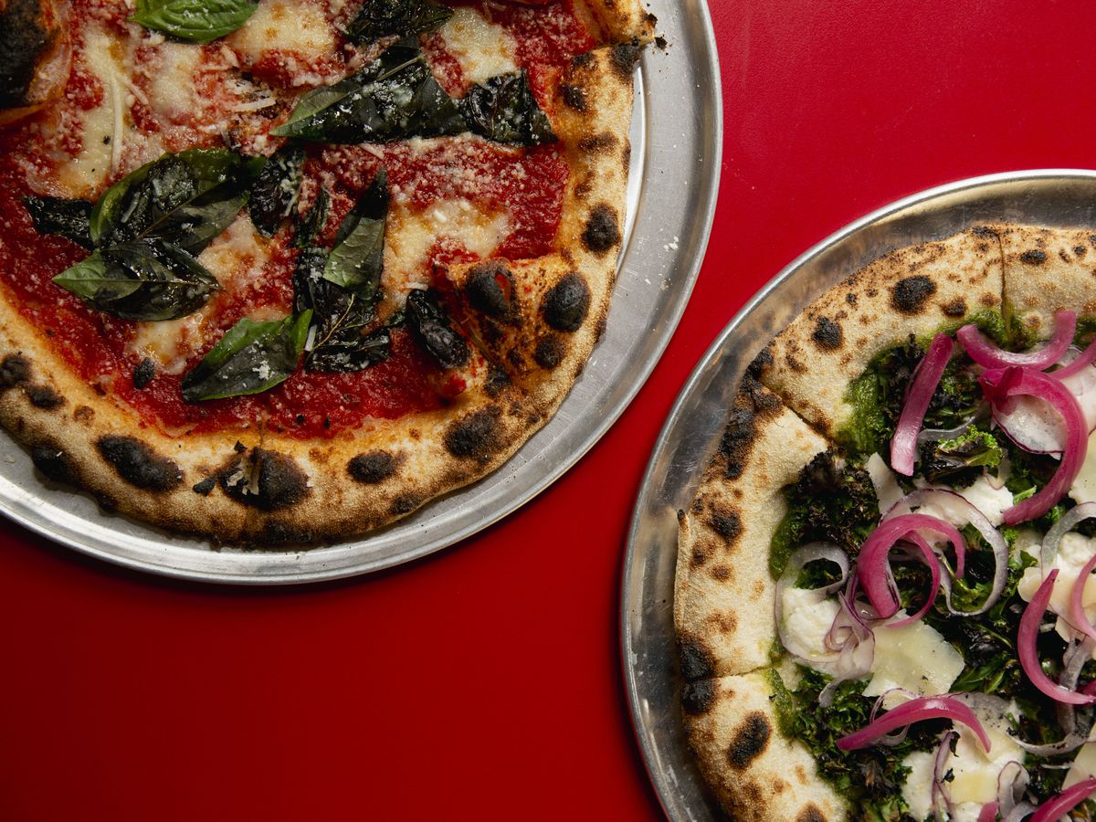 Two pizzas on trays on a red table.