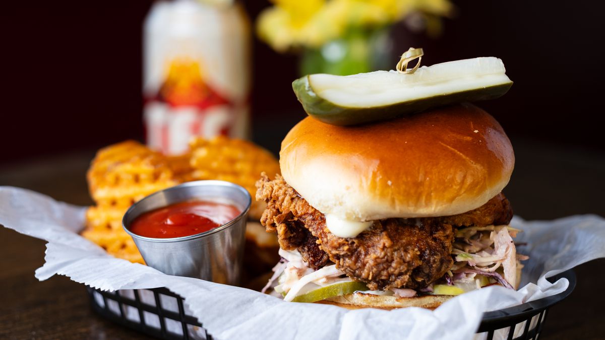 A chicken sandwich on a bun sits in a black basket with a pickle on top, held in place with a toothpick. Also in the basket are waffles fries and a ramekin of ketchup.