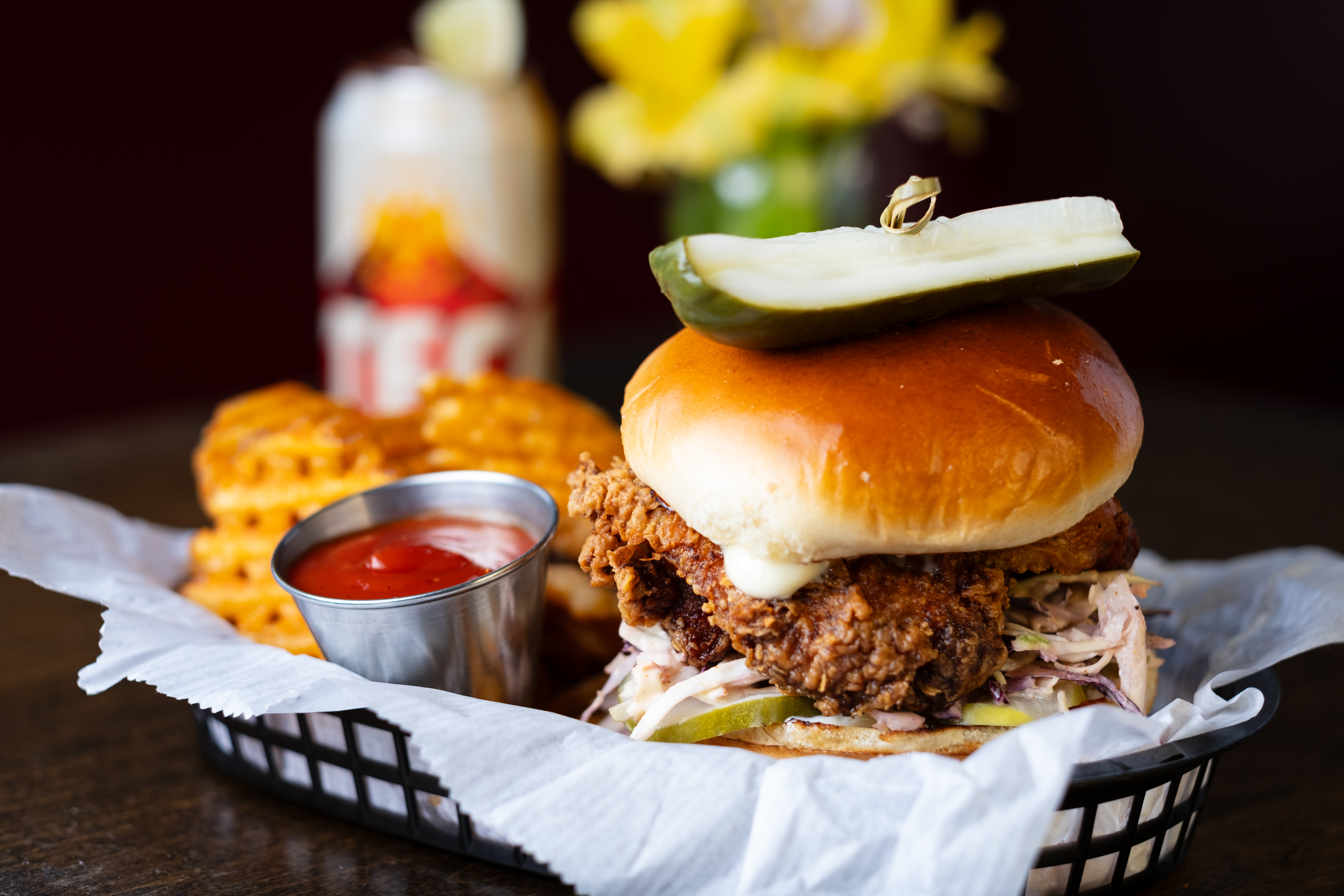 A chicken sandwich on a bun sits in a black basket with a pickle on top, held in place with a toothpick. Also in the basket are waffles fries and a ramekin of ketchup.