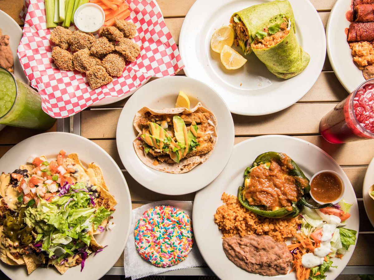 A spread of vegetarian dishes includes (clockwise) a burrito, faux chicken tacos, enchiladas with red sauce, a stuffed bell pepper, a cookie with sprinkles, nachos, and fried zucchini.