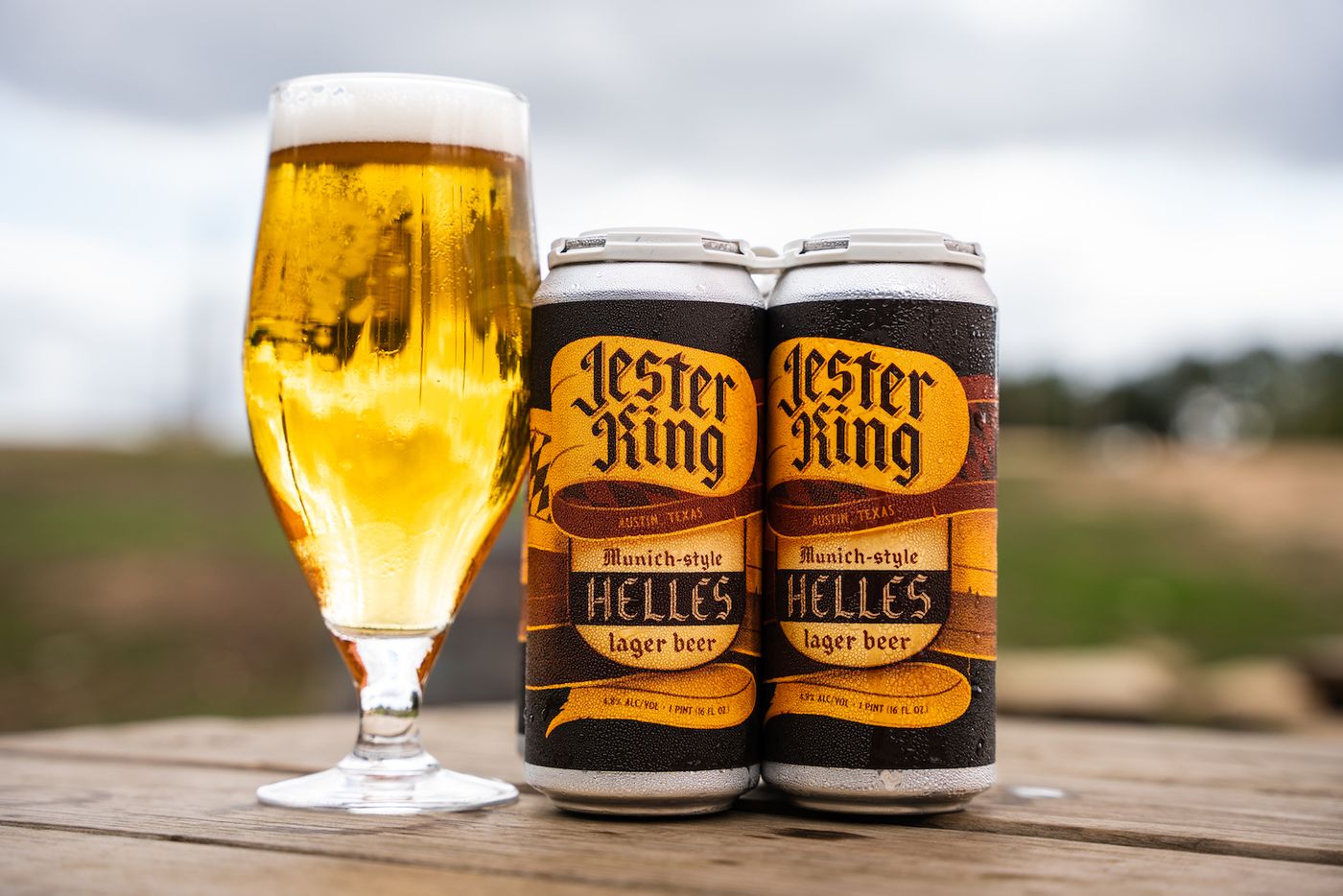 A tall glass of beer next to two cans of beer.