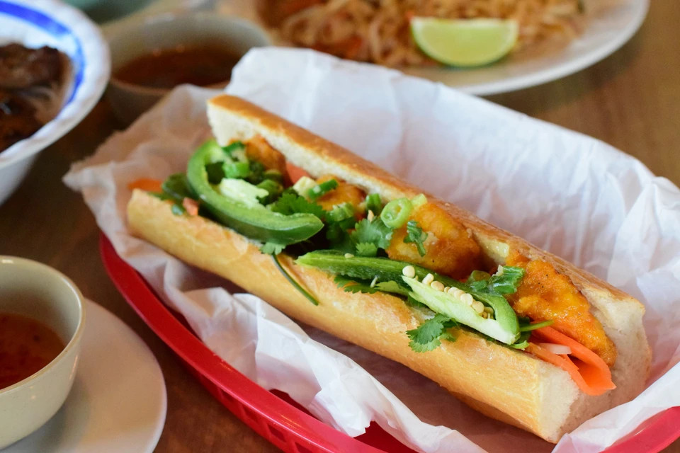 A banh mi stuffed with fried tofu, shredded carrots, and peppers sits in a red boat lined with white paper.