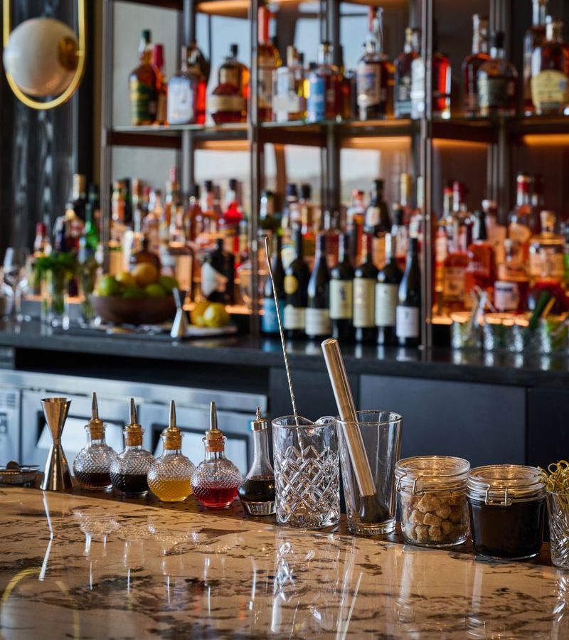 A bar is visible, with bottles shelved in the background and mixers and bitters lined up on the bar.