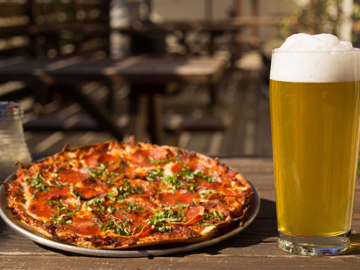 A pepperoni pizza on a silver tray sites on a table next to a giant glass of beer with a foamy head.