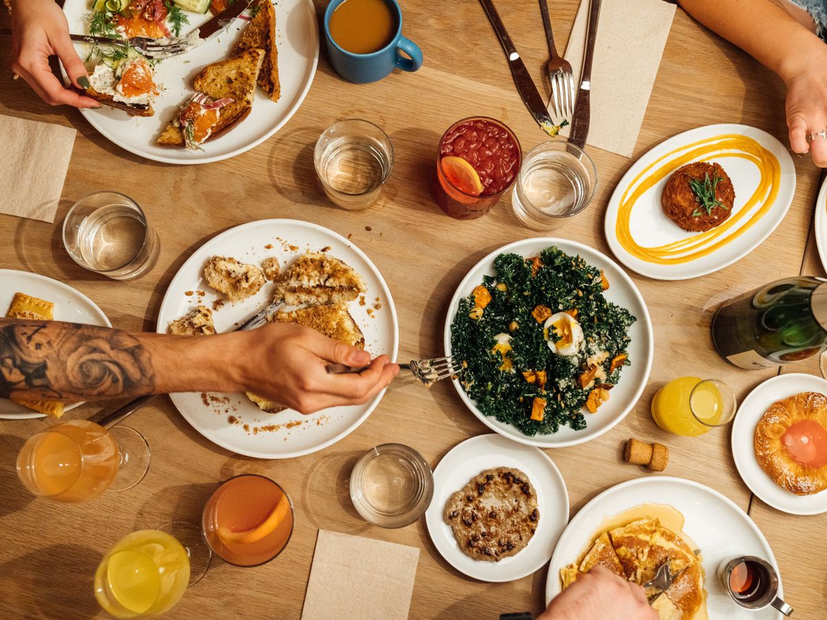 A table holds various brunch dishes. Clockwise: a hash brown, a fruit danish, a plate of greens with some orange squash, pancakes, oatmeal, fried fish, and eggs and toast.