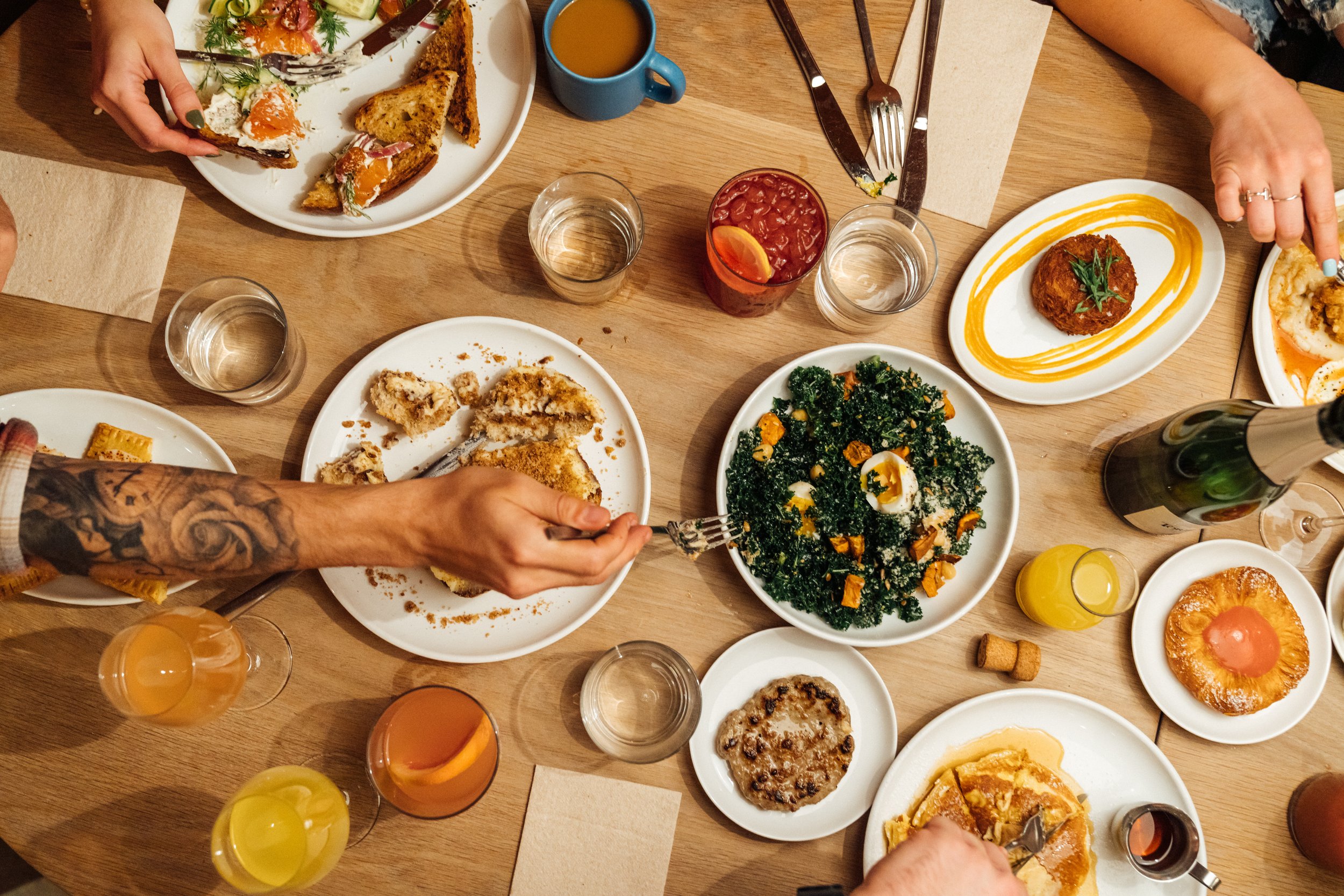 A table holds various brunch dishes. Clockwise: a hash brown, a fruit danish, a plate of greens with some orange squash, pancakes, oatmeal, fried fish, and eggs and toast.