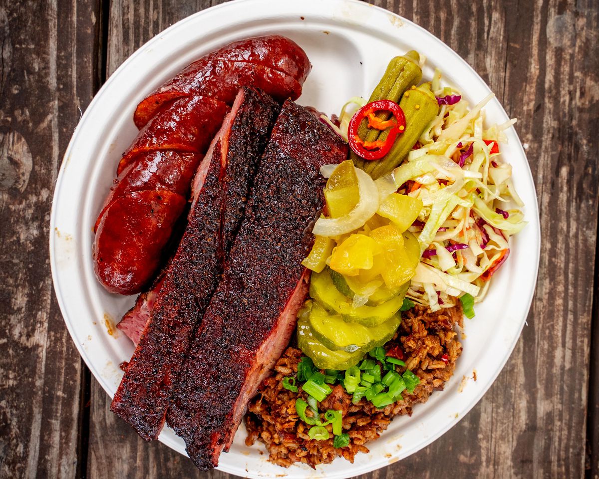 A plate of barbecue has a slice of fatty brisket, sausage, dirty rice, pickled vegetables, and cole slaw.