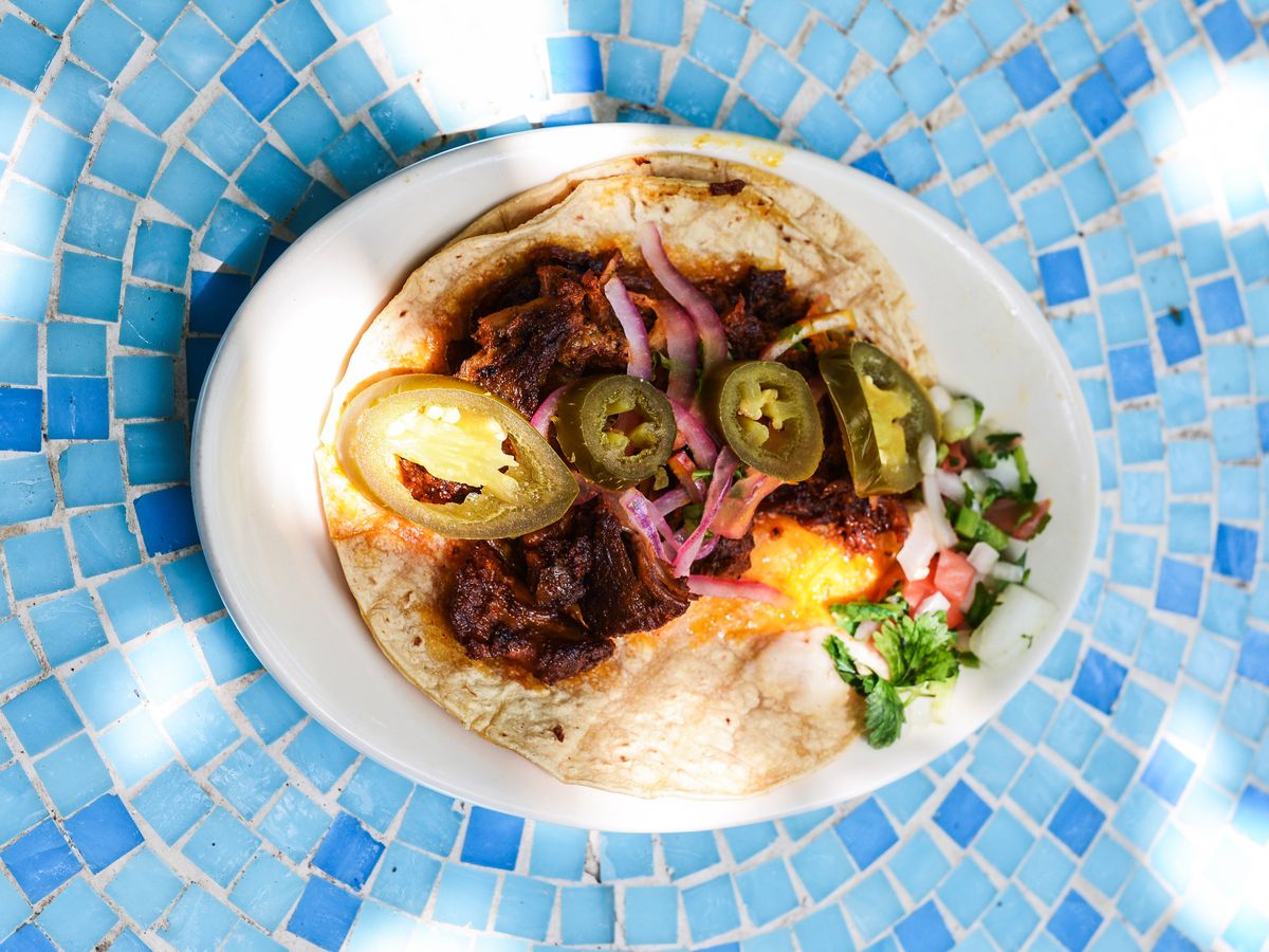 An oval white plate with a pale brown tortilla topped with brown meat, green jalapeño slices, and pickled onions.