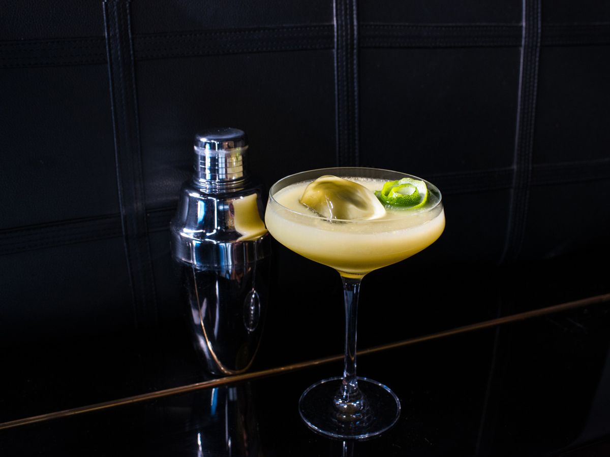 A cloudy yellow cocktail in a daiquiri glass against a very black room.