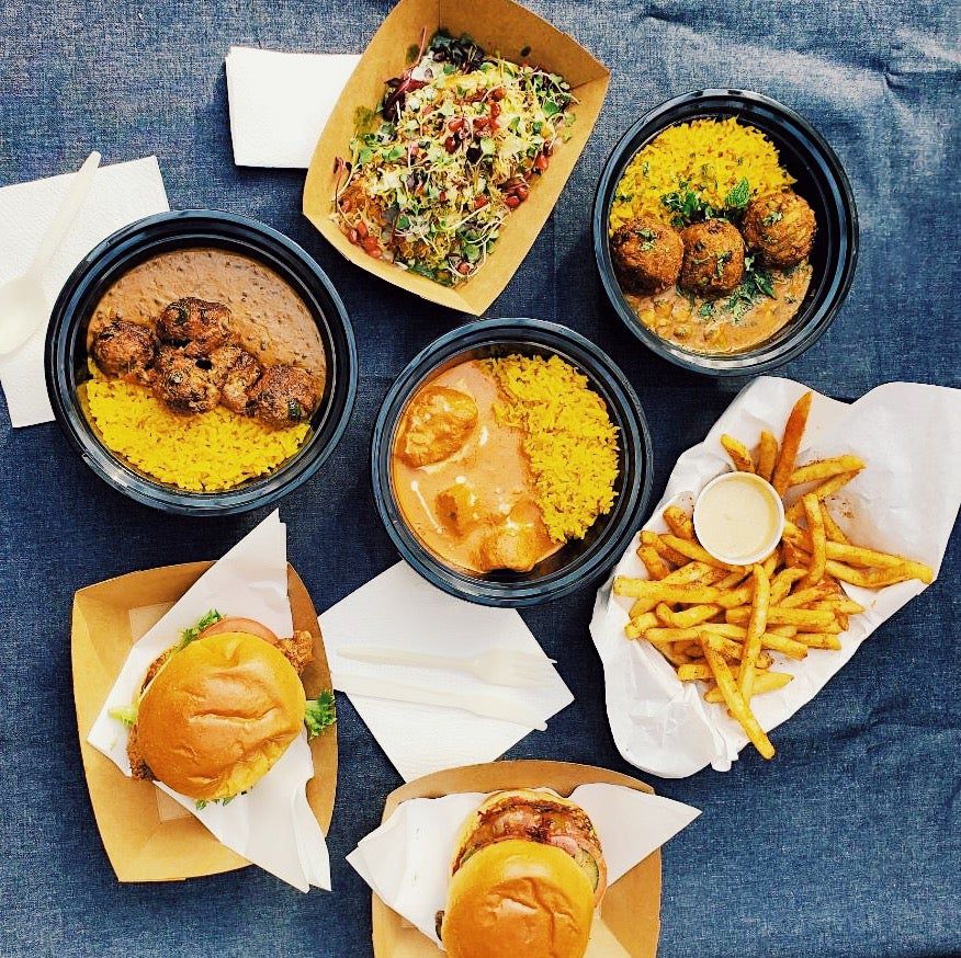 Several dishes sit together on a table, including (clockwise) a salad, chicken curry, meatballs, french fries, a burger, a second burger, and roti with rice and beans.