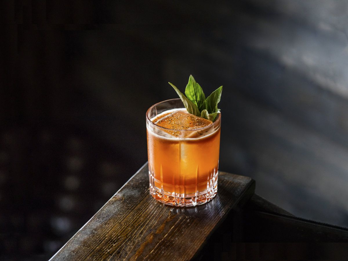 A brown-ish orange cocktail sits on the edge of a table, garnished with mint.